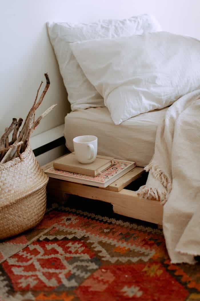services-01 Comfortable cozy bedroom with bed wooden shelves with book and cup while wicker basket with sticks placed on ethnic styled rug