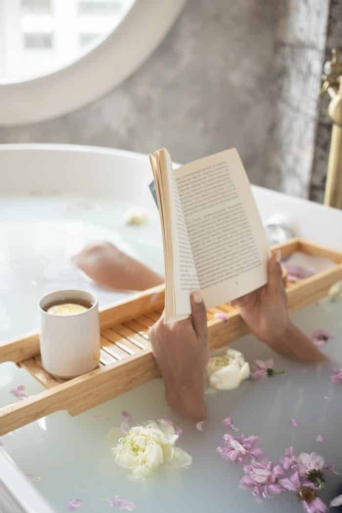 approach-img From above of unrecognizable female lying in bathtub full of water with delicate ivory and pink flowers and reading book on blurred background